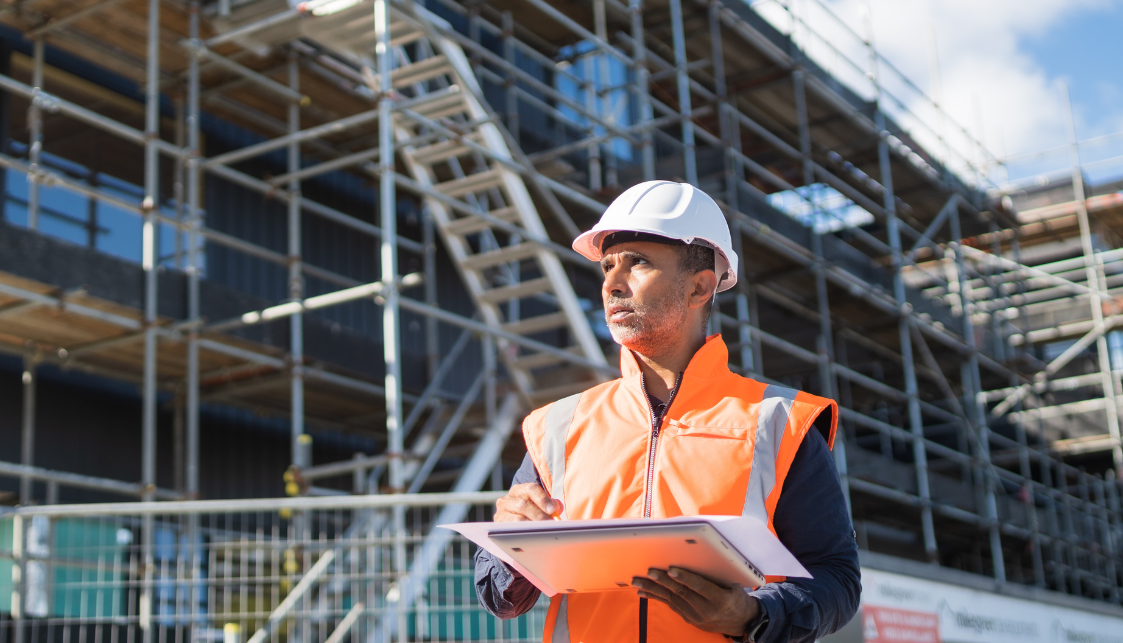 Un chef de chantier avec un gilet orange et un casque, devant un immeuble en construction, et tenant dans ses mains une tablette, un plan et un crayon.