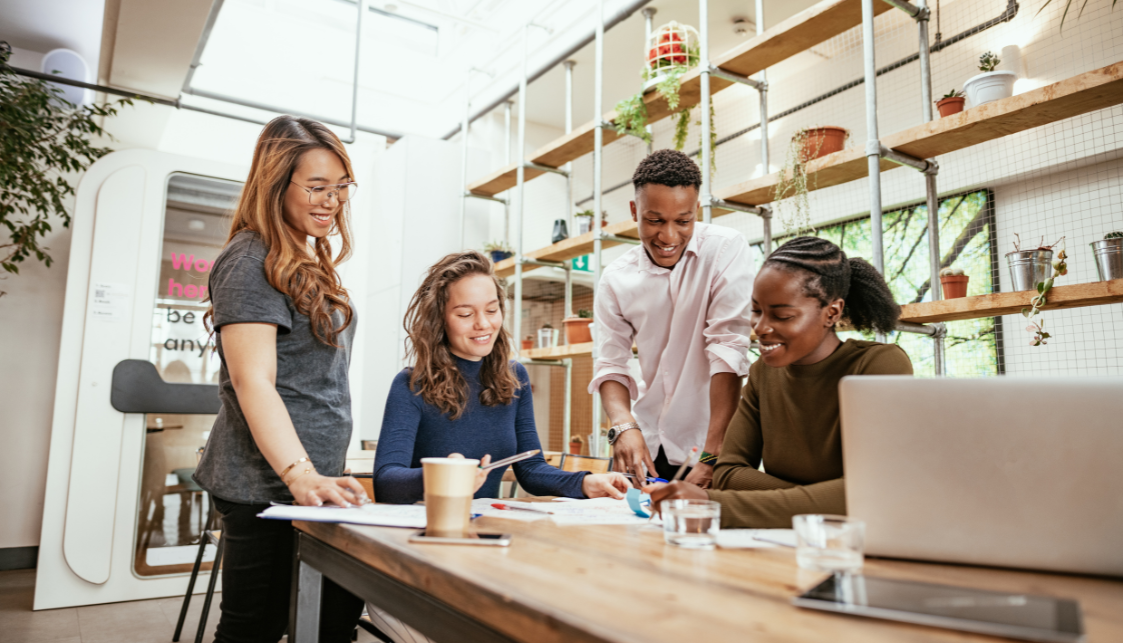 4 personnes, 3 femmes et un homme, dans une salle lumineuse au tour d'une table, 2 sont debouts, 2 sont assises. Sur la table il y a un PC portable au premier plan, des petits verres d'eau, un gobelet de café, et des documents que sont en train de regarder les personnes. le mur derrière eux est habillé d'étagères en bois et armature metallique.