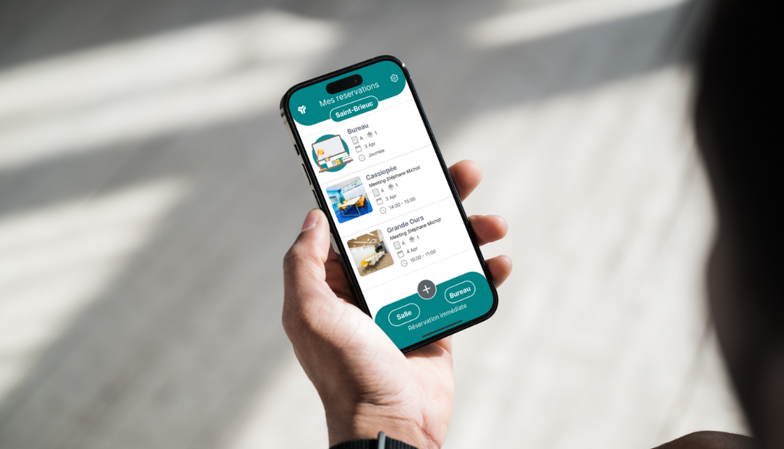 Un smartphone dans une main avec sur l'écran une liste de réservation de salles et de bureau.