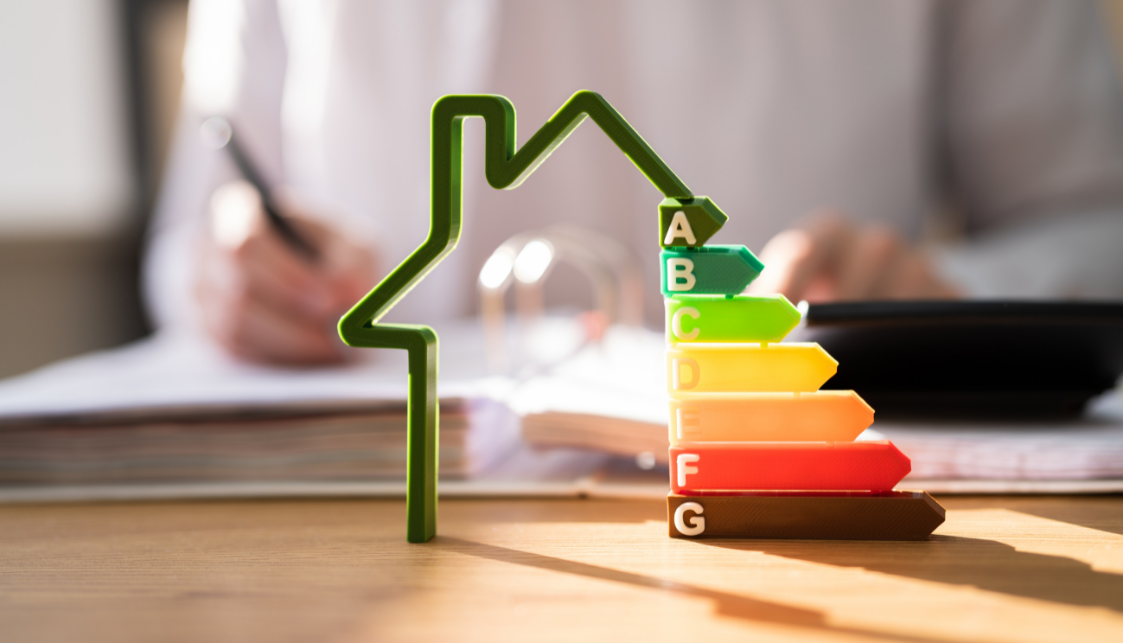 Au premier plan, sur une table, un objet ouvert représentant une maison et les 6 catégories de consommation d'énergie de A à G réprésentées par des flèches en dégradé de couleur allant du vert au rouge. En arrière plan, flou, une personne assise à son bureau écrivant sur un classeur épais.