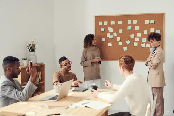 employees sitting around a table brainstorming with an employee in front of a board where post-it notes are stuck