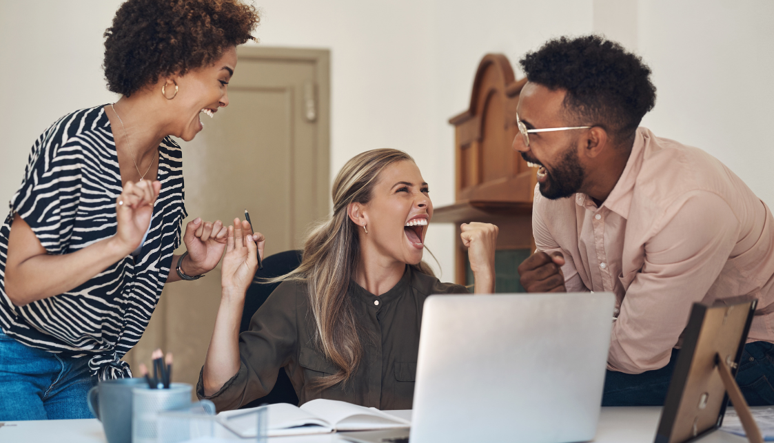 Trois personnes riant ensemble dont une au milieu assise à son bureau avec son ordinateur portable devant elle