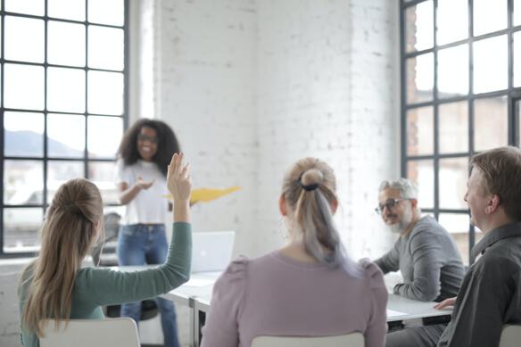 a collaborator raise his hand, they are four sitting around a table and listen to a woman standing in front of them