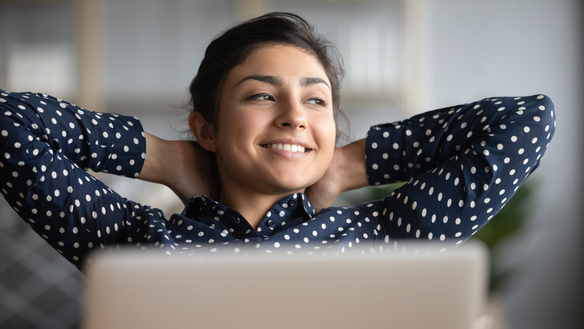 A smiling woman sitting in front of her laptop, arms crossed behind her head.