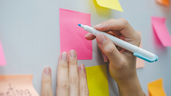 Des post-it de couleur collés sur un mur. Une main qui tient un feutre bleu et qui s'apprête à écrire sur l'un des post-it, avec l'autre main qui tient le post-it.