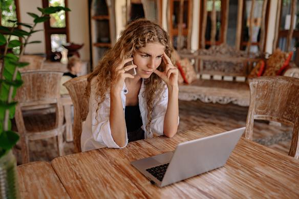 a woman sitting at her dining room table in front on a laptop and her mobile phone in her ear, looking worried